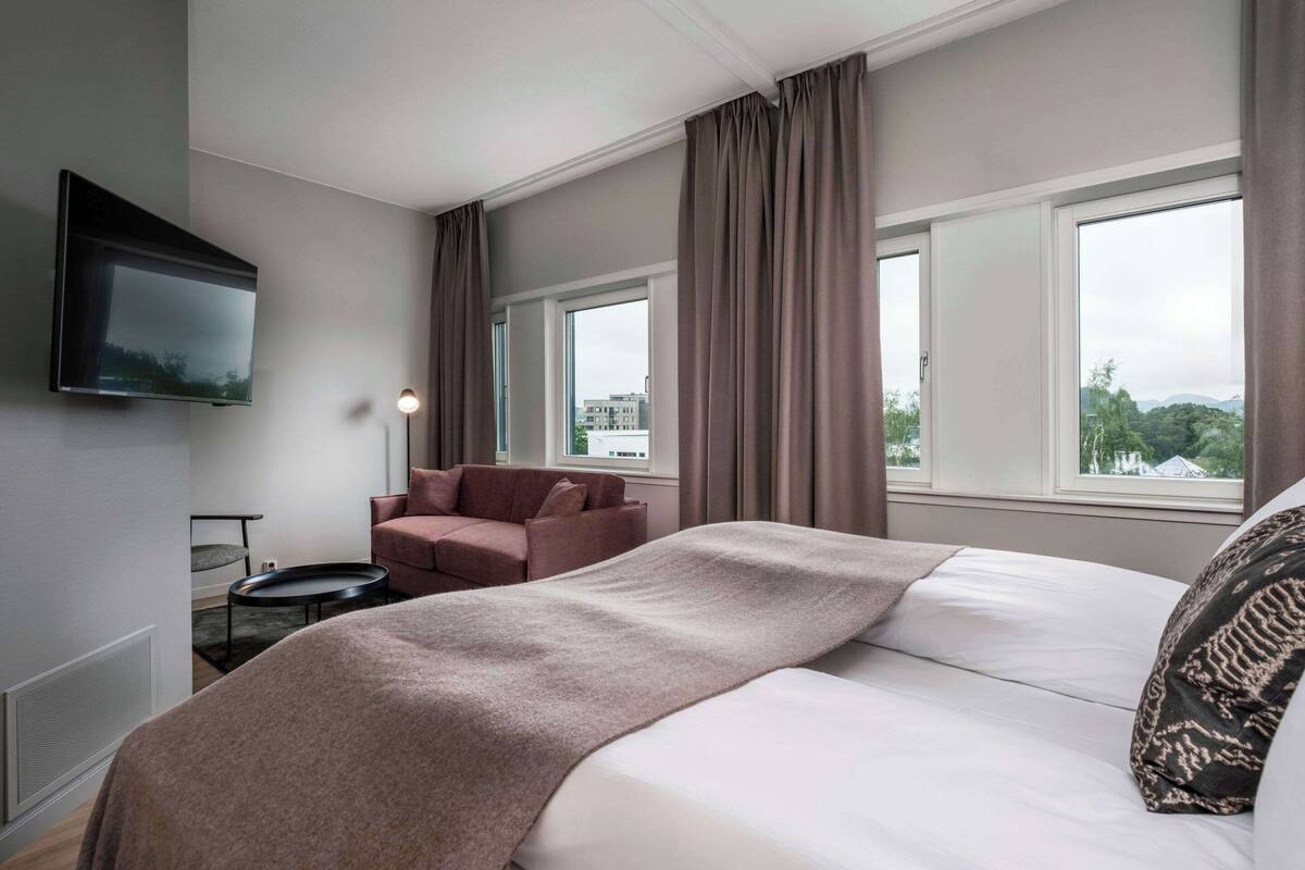 Ein modernes Hotelzimmer mit einem großen Bett, einem Flachbildfernseher, einer Couch und Blick auf die Stadt durch große Fenster.
