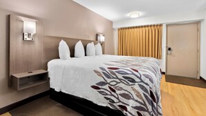 A hotel room with a large bed, a bedside table, a wall-mounted light, and a door with curtains. - Red Roof Inn Harrisburg North (Harrisburg)