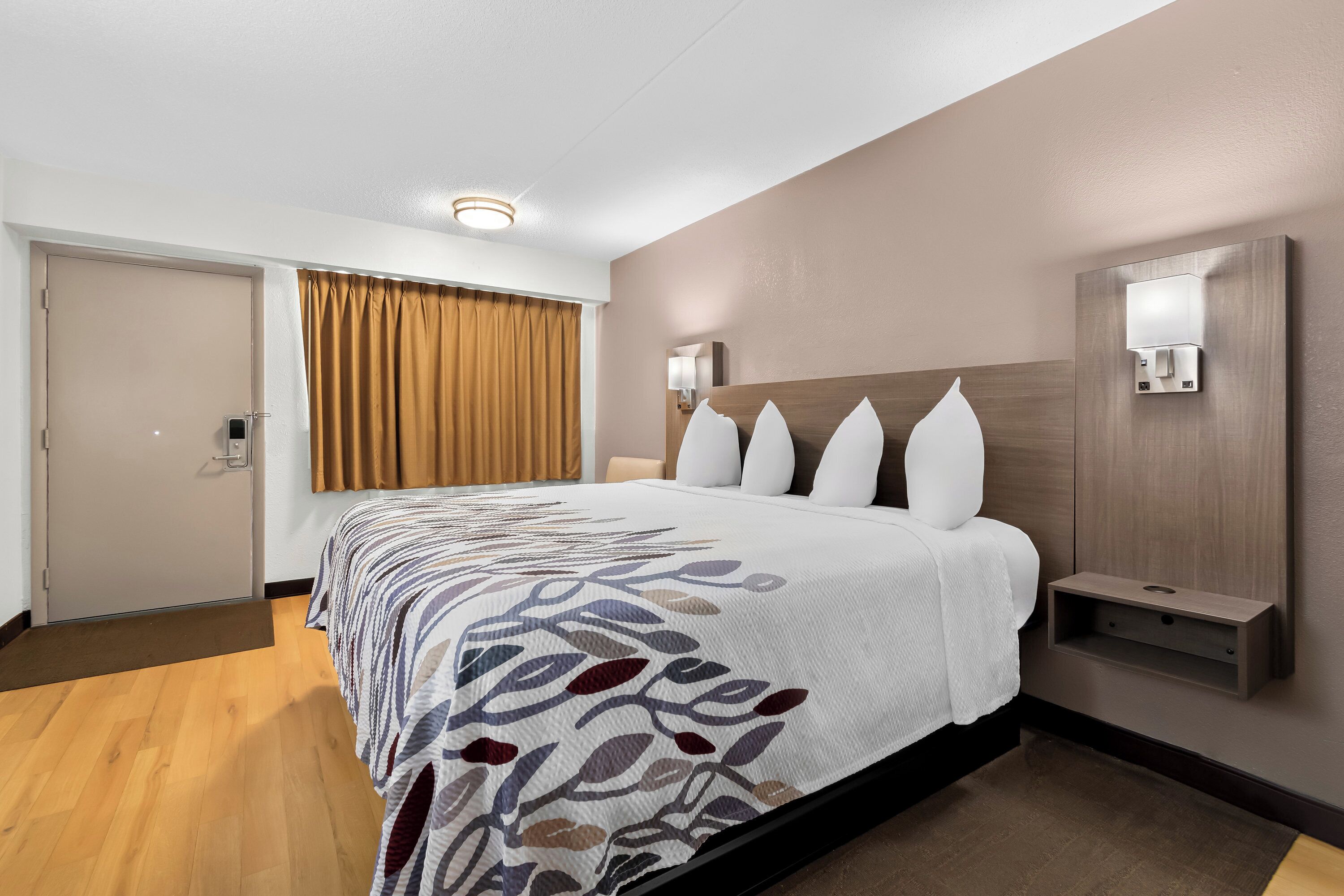 A hotel room with a large bed, a bedside table, a wall-mounted lamp, and a door with a curtain.