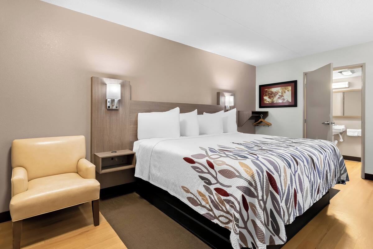Une chambre d’hôtel avec un lit, une chaise, une table de chevet, une lampe fixée au mur et un tableau accroché au mur.