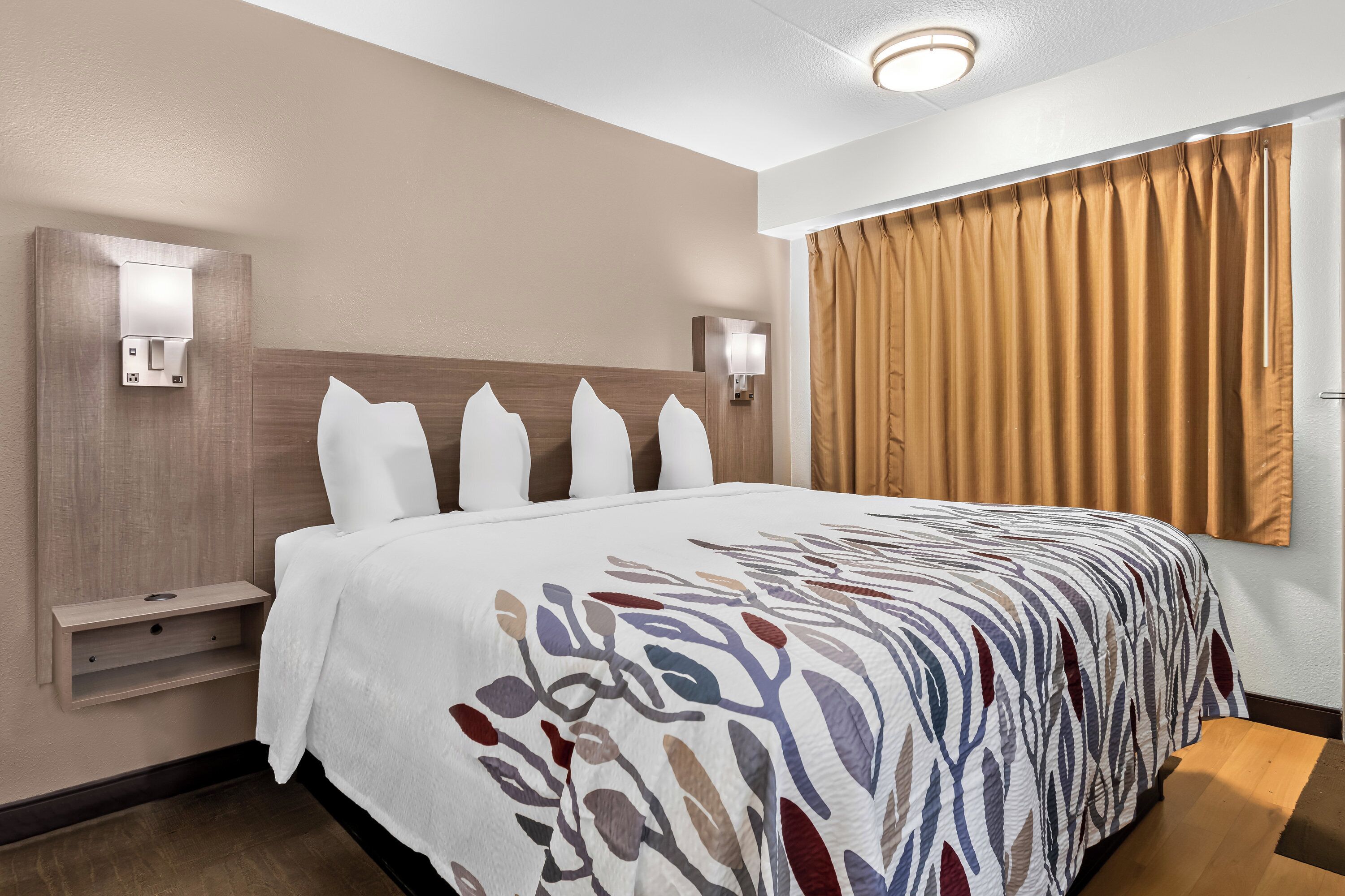 A hotel room with a large bed, a bedside table, a wall-mounted light, and a window with curtains.