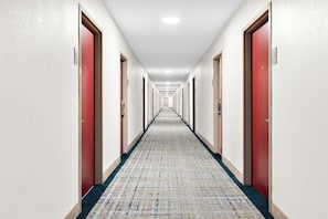 Een lange, smalle hotelgang met rode deuren aan beide kanten en een kamerbreed tapijt met dessin.