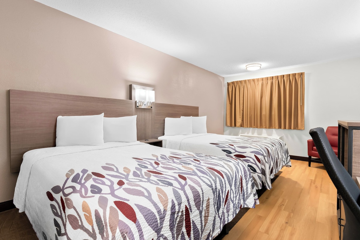 Ein Hotelzimmer mit zwei Betten, einem Schreibtisch, einem Stuhl und einem Fenster mit Vorhängen.