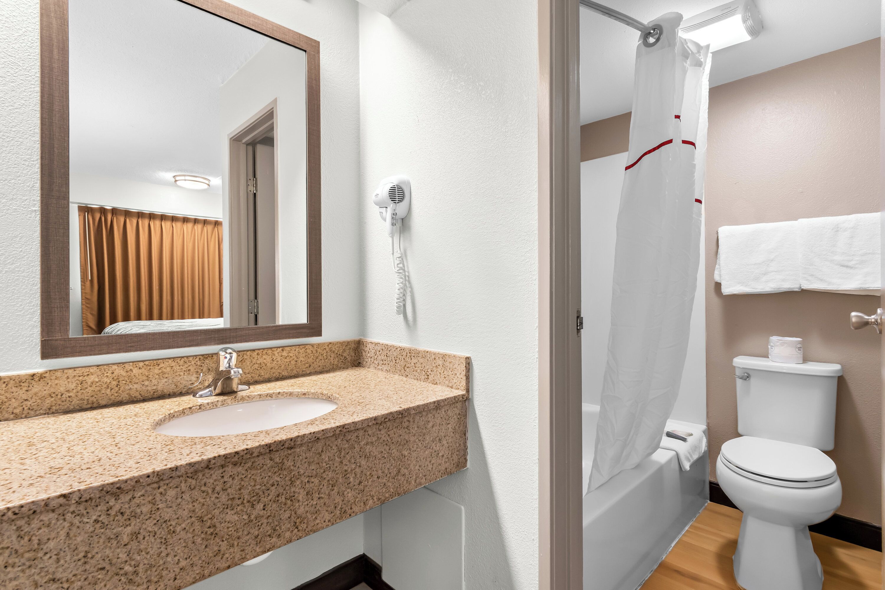 Une salle de bain équipée d’un lavabo avec un plan de travail en granit, d’un miroir, d’un sèche-cheveux, d’un rideau de douche, de toilettes et d’un porte-serviettes.
