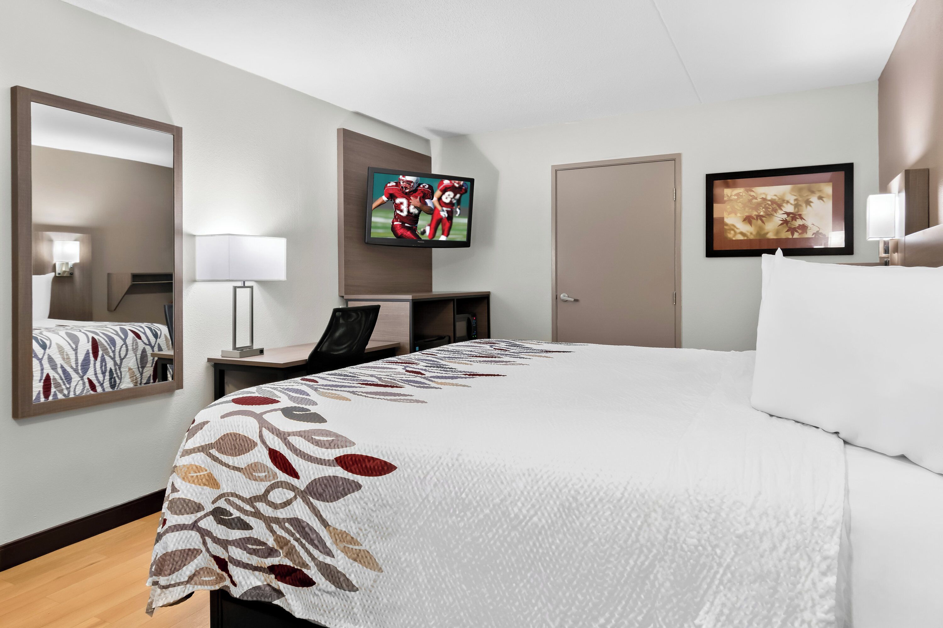 A hotel room with a large bed, a desk, a TV mounted on the wall, and a door.