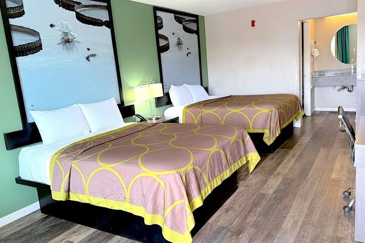 Ein Hotelzimmer mit zwei Betten, einem Schreibtisch, einem Spiegel und einem durchgehenden Türrahmen sichtbaren Badezimmer.