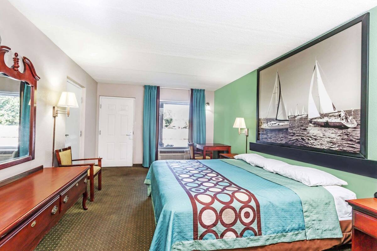 Ein Hotelzimmer mit Bett, Schreibtisch, Stuhl, Kommode, einem Fenster mit Vorhängen und einem Gemälde mit Segelbooten an der Wand.