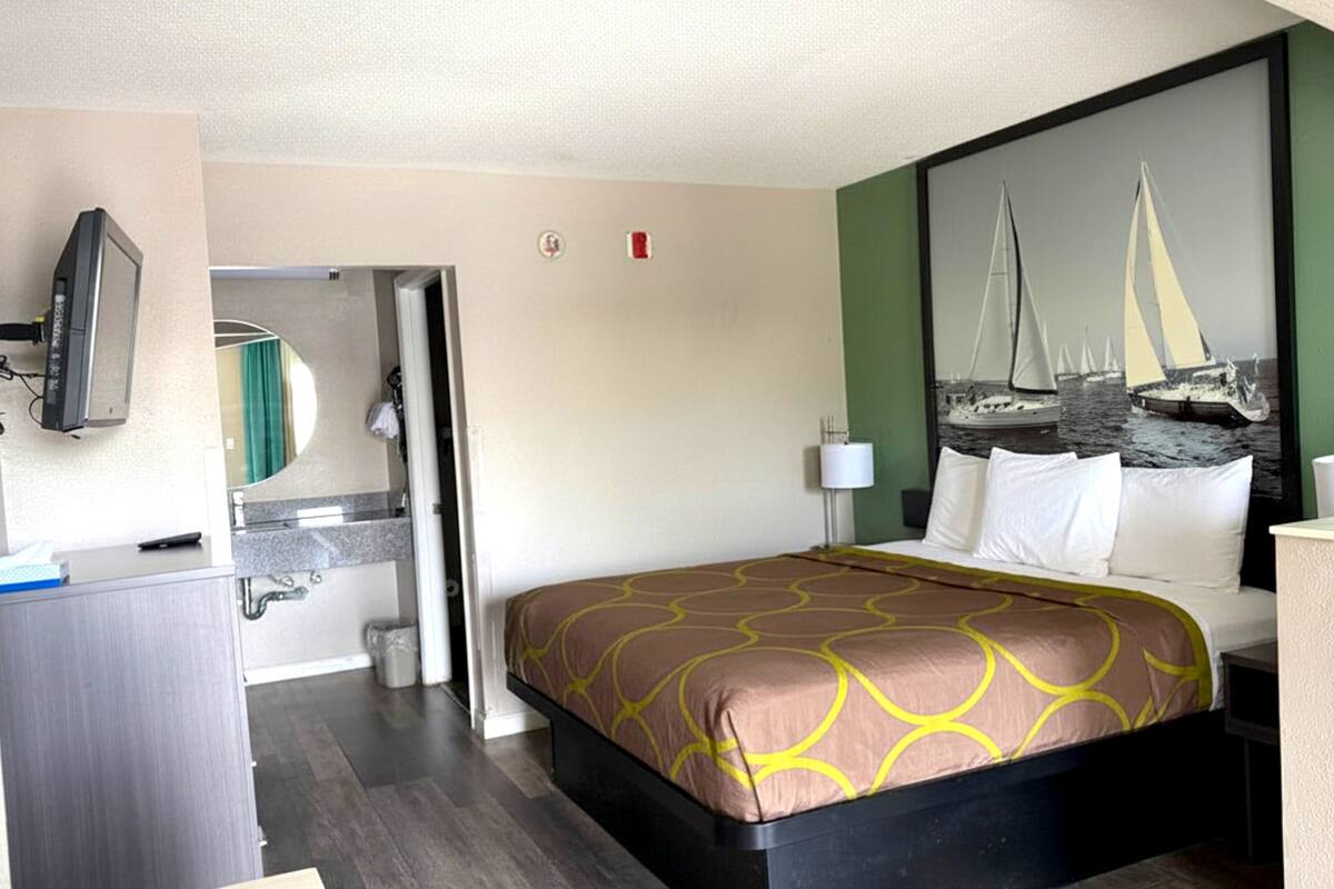 Ein Hotelzimmer mit einem Bett, einem Schreibtisch, einem Spiegel, einem Bild von Segelbooten und einem durch eine offene Tür einsehbaren Badezimmer.