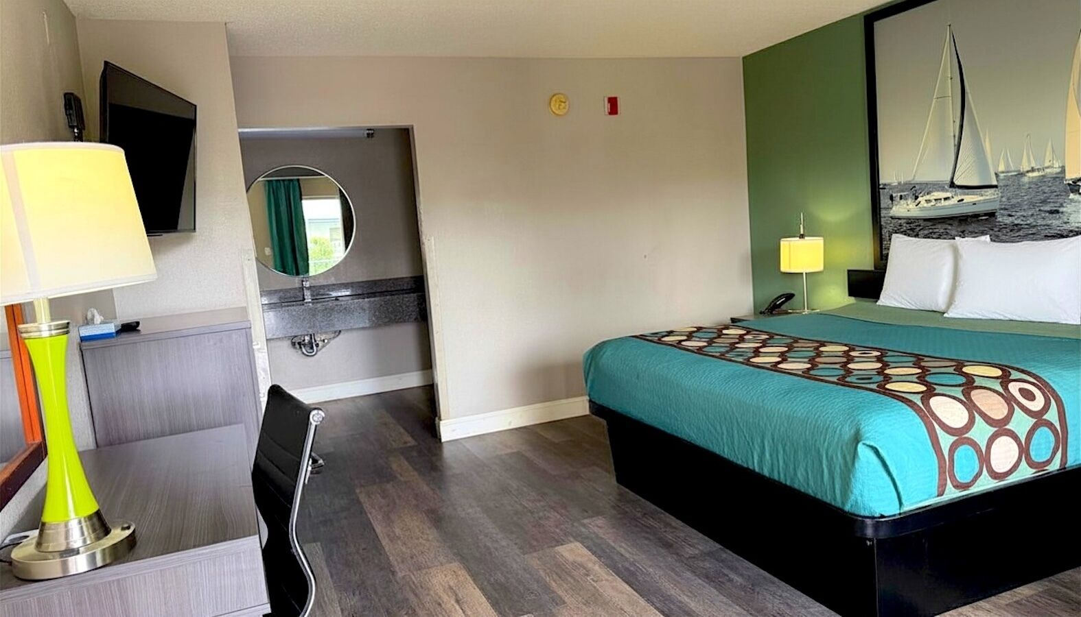 A hotel room with a bed, a desk, a chair, a mirror, a television, and a painting on the wall.