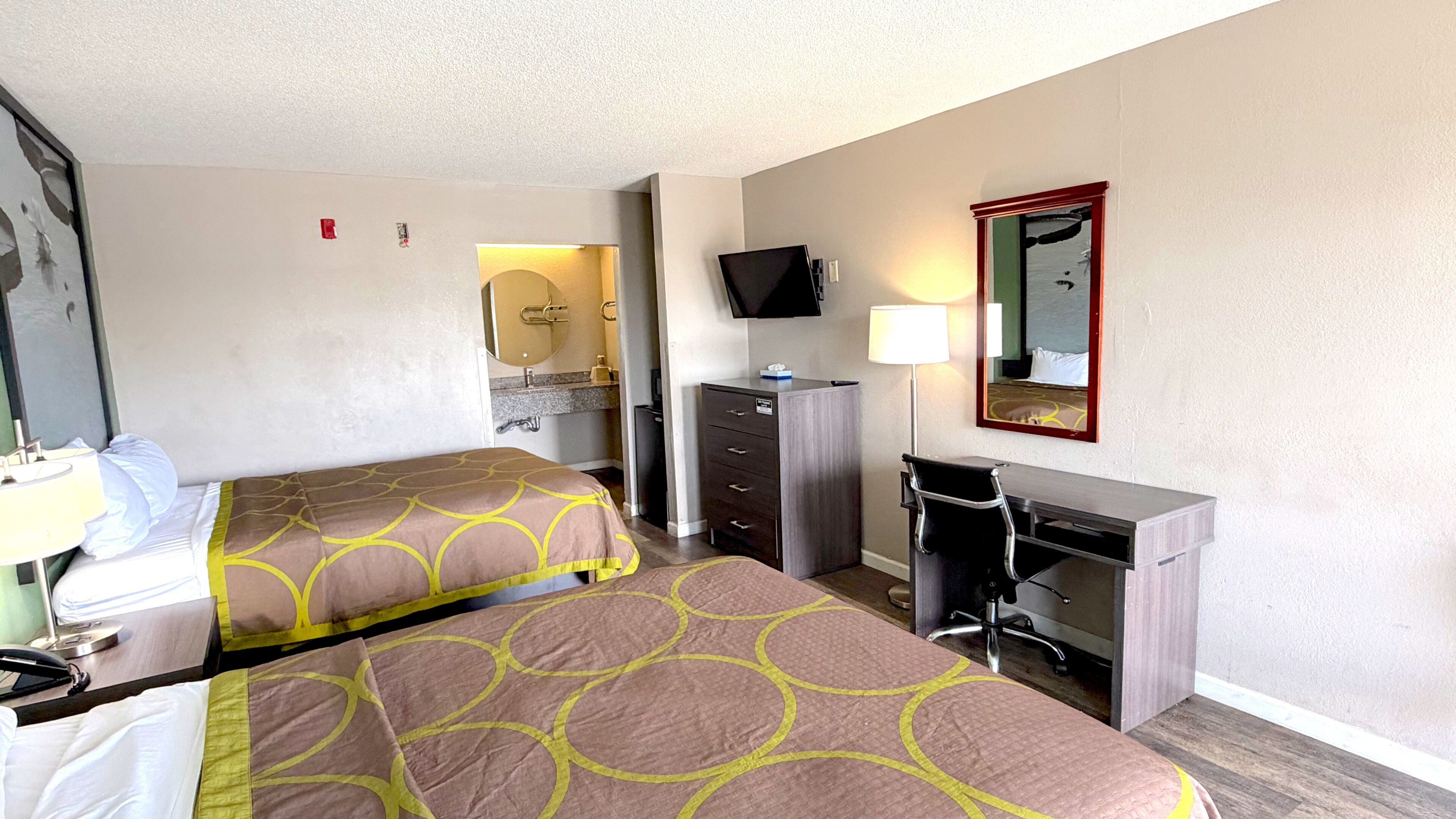 A hotel room with two beds, a desk, a chair, a TV, a mirror, and a bathroom visible through an open door.