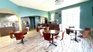 Un comedor con mesas y sillas redondas, una cocina al fondo y un ventanal grande.