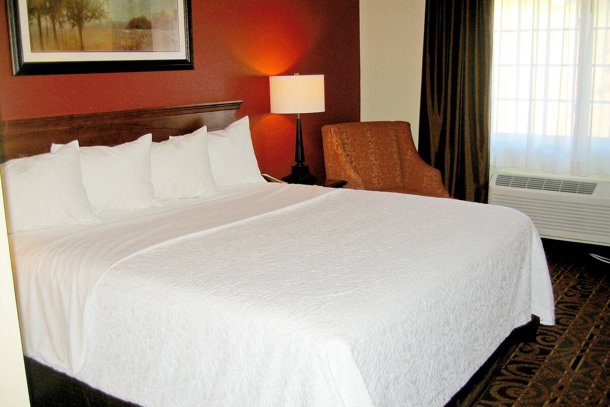 Habitación de hotel con una cama grande, una mesita de noche, una lámpara, una silla y un ventanal con cortinas.