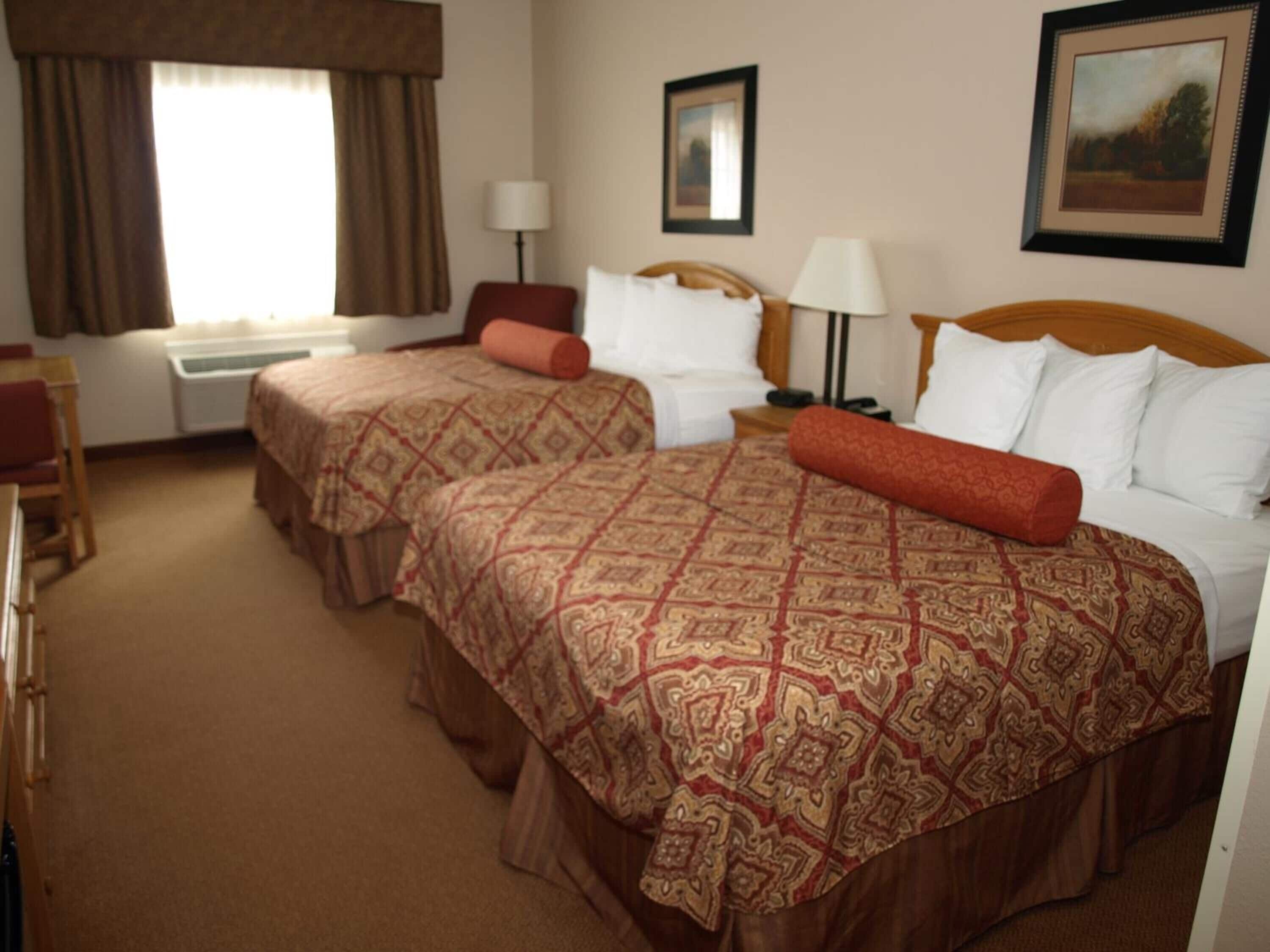 A hotel room with two beds, a desk, a chair, a window with curtains, a lamp, and a framed picture on the wall.