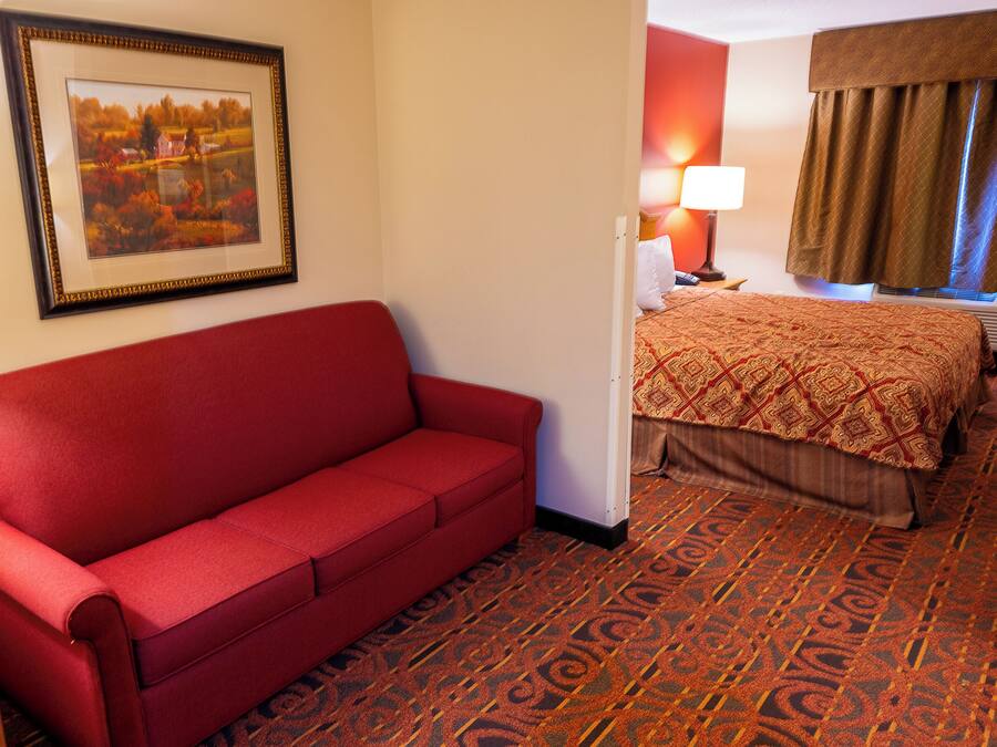 A hotel room with a red sofa, a framed picture on the wall, a bed with a patterned bedspread, and a lamp on the nightstand.