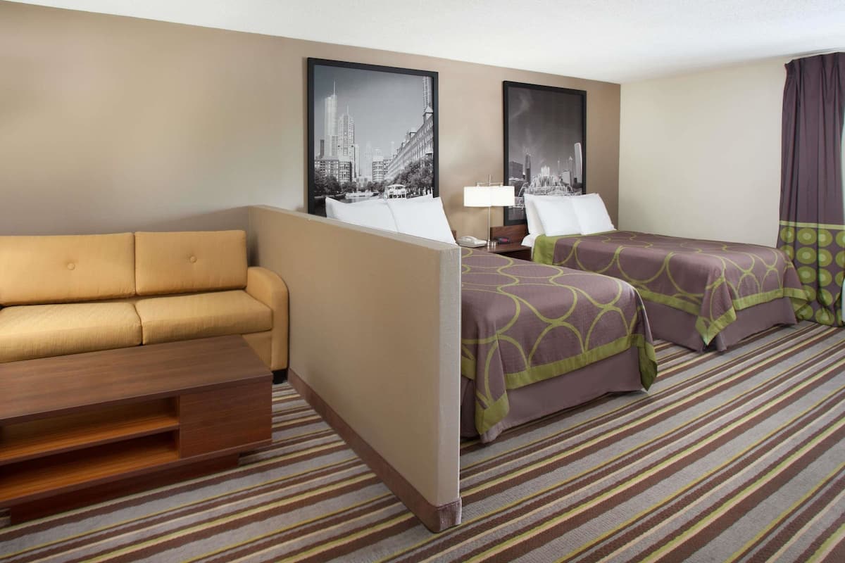 A hotel room with a sofa, two beds with patterned bedspreads, a nightstand, a lamp, and two framed pictures on the wall.
