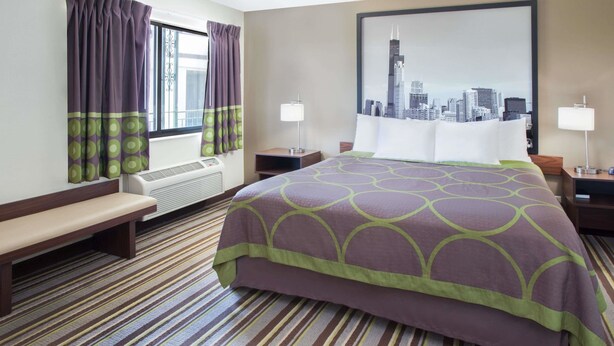 A hotel room with a bed, bench, air conditioning unit, two bedside tables with lamps, and a cityscape painting on the wall. - Super 8 by Wyndham Chicago/Rosemont/O'Hare/SE (River Grove)