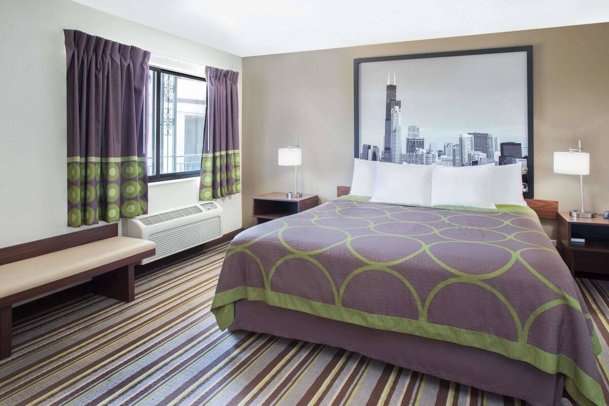 A hotel room with a bed, bench, air conditioning unit, two bedside tables with lamps, and a cityscape painting on the wall.