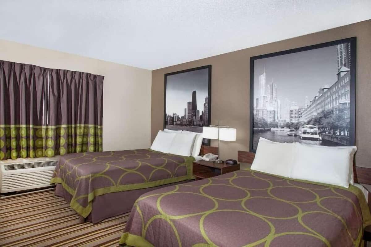 A hotel room with two beds, a large window with curtains, a cityscape painting, and a nightstand with a lamp.