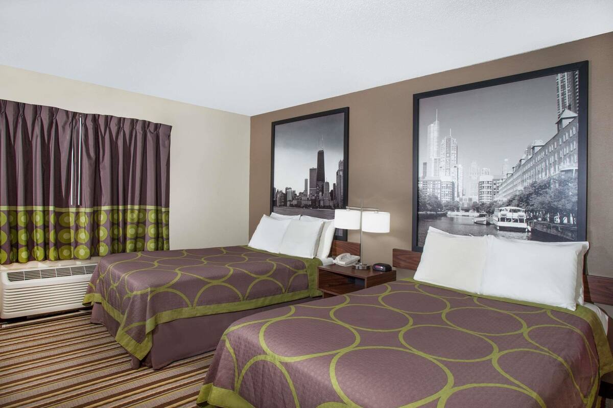 A hotel room with two beds, a large window with curtains, a cityscape painting on the wall, and a flat-screen TV.