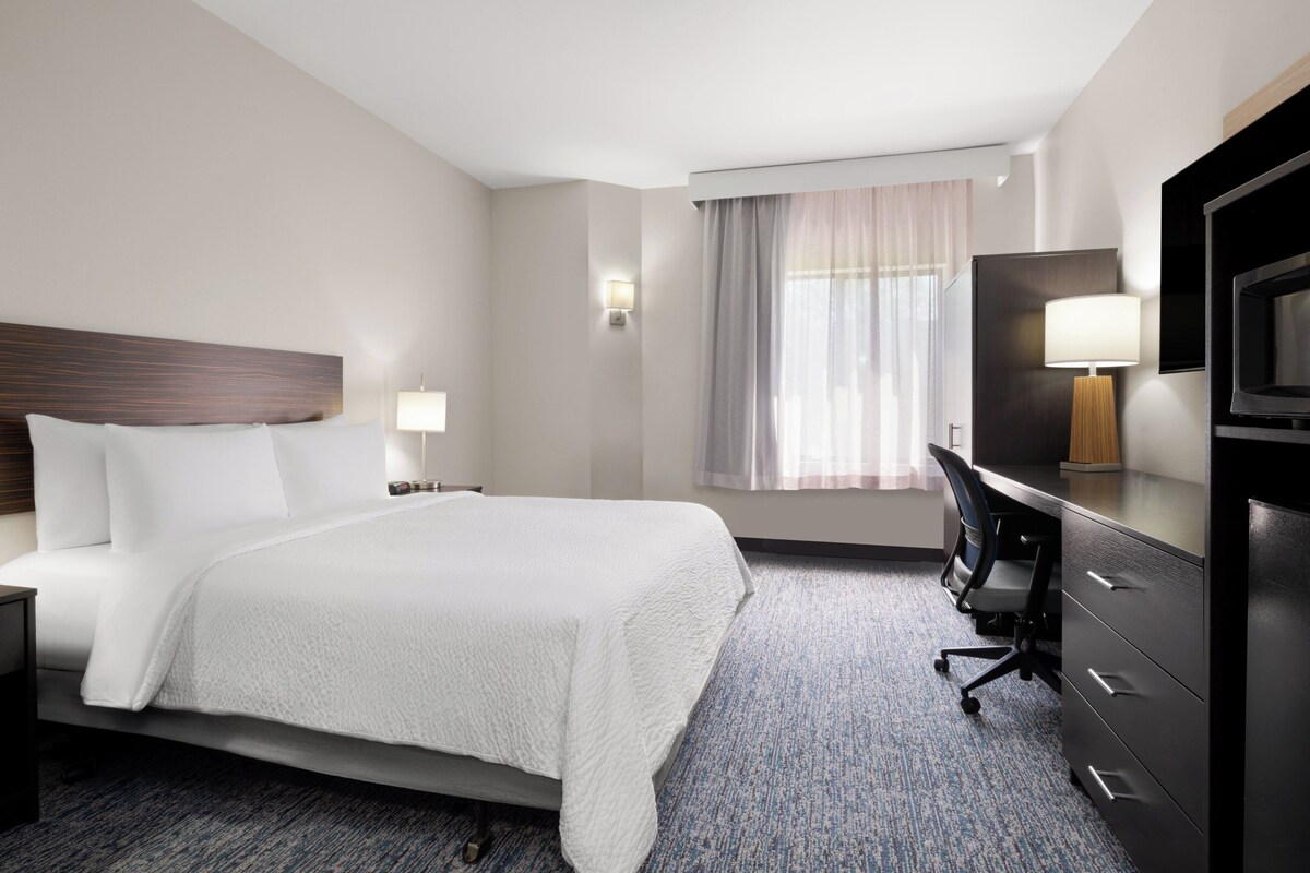 Quarto de hotel com uma cama grande, uma escrivaninha com cadeira, televisão e janela com cortinas.