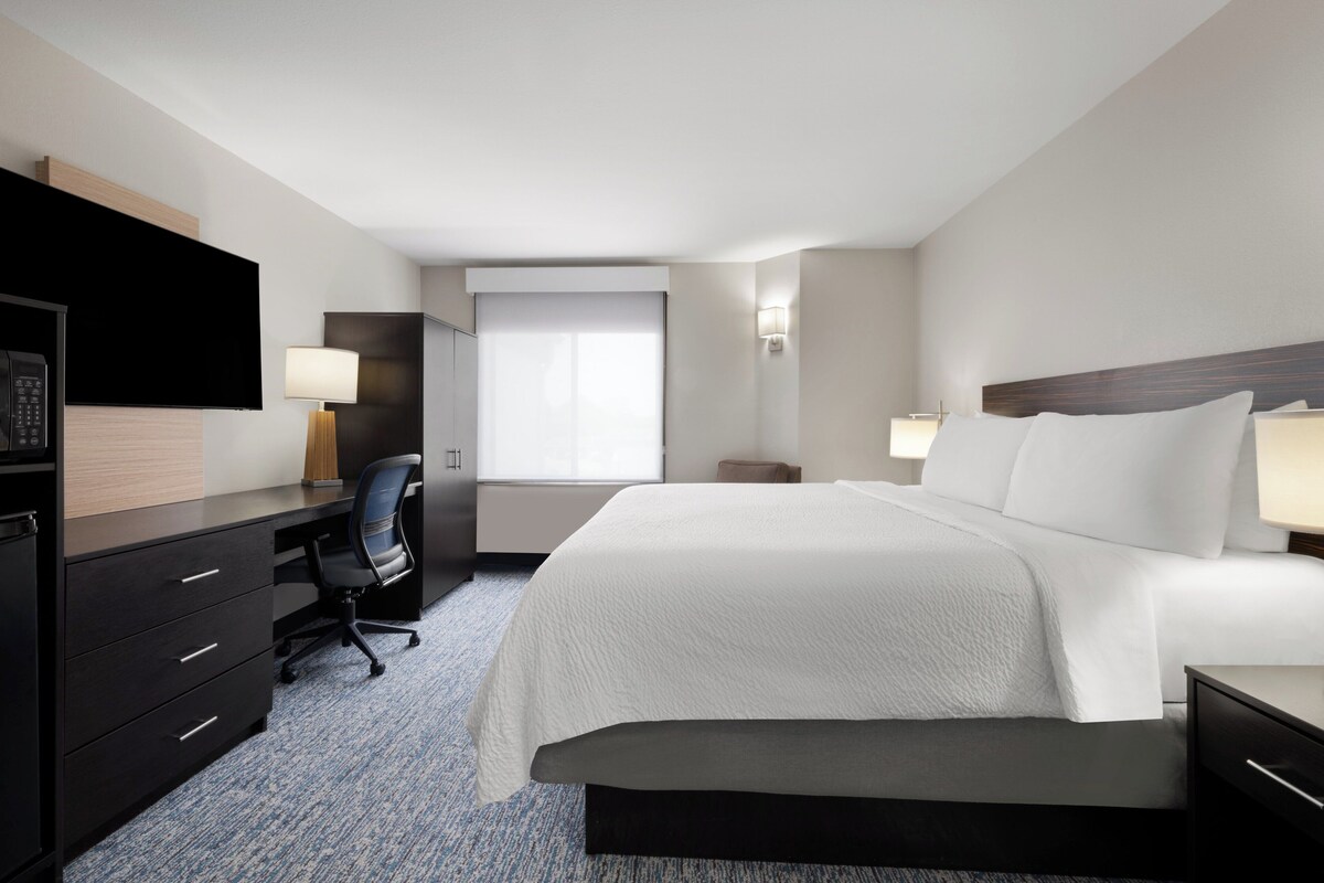 Quarto de hotel com uma cama grande, escrivaninha, cadeira, televisão e janela com cortinas.