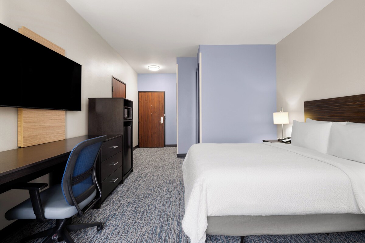 Quarto de hotel com cama, escrivaninha, cadeira, televisão e porta.
