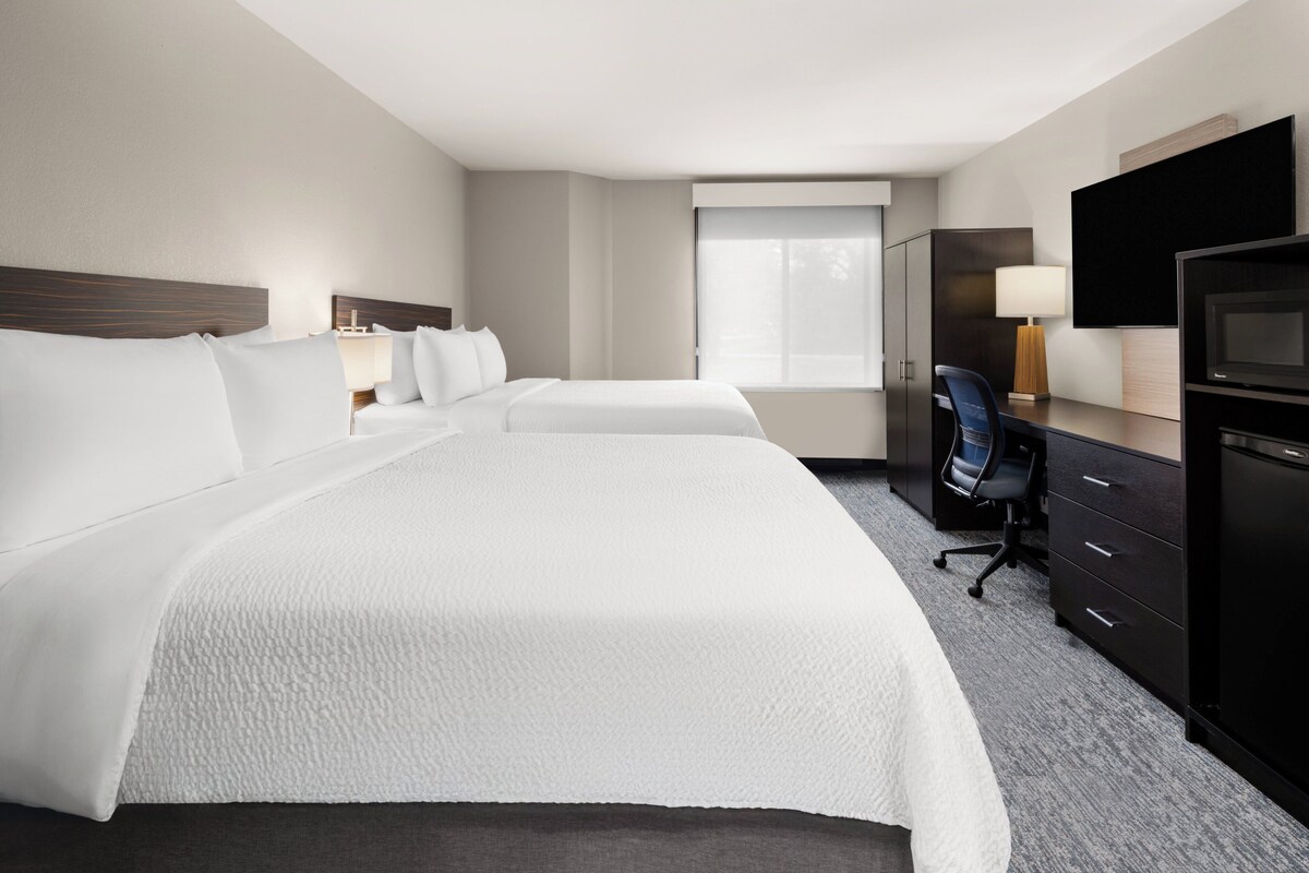 Quarto de hotel com duas camas, uma escrivaninha, uma cadeira, uma televisão e um guarda-roupa.
