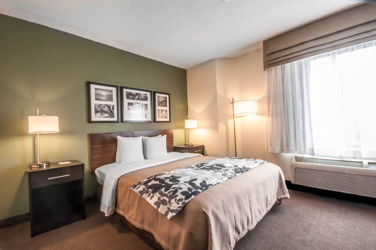 A hotel room with a bed, two bedside lamps, a nightstand, a wall-mounted air conditioner, and framed pictures on the wall.