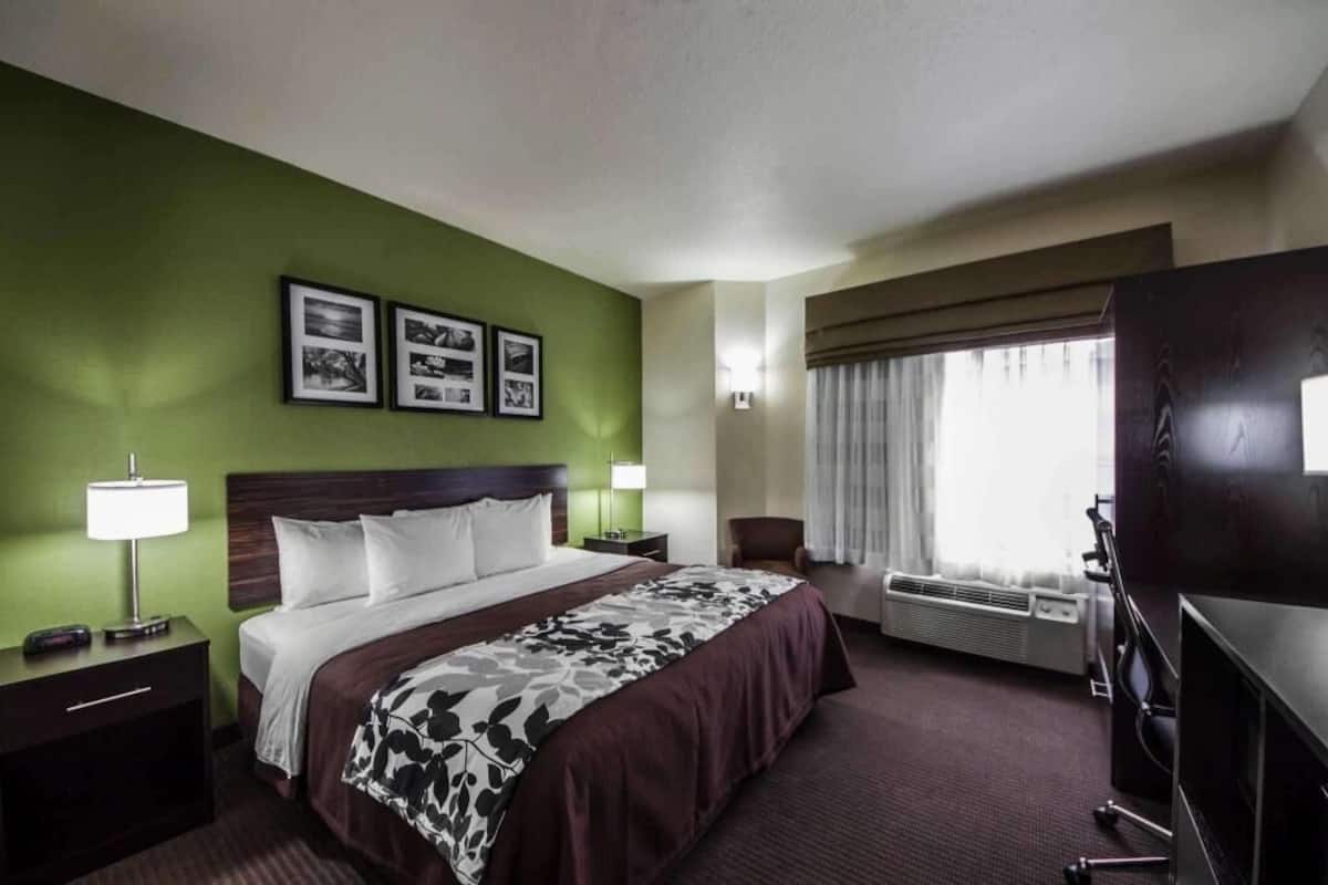 A hotel room with a bed, two bedside tables, a TV, a chair, a window with curtains, and framed pictures on the wall.