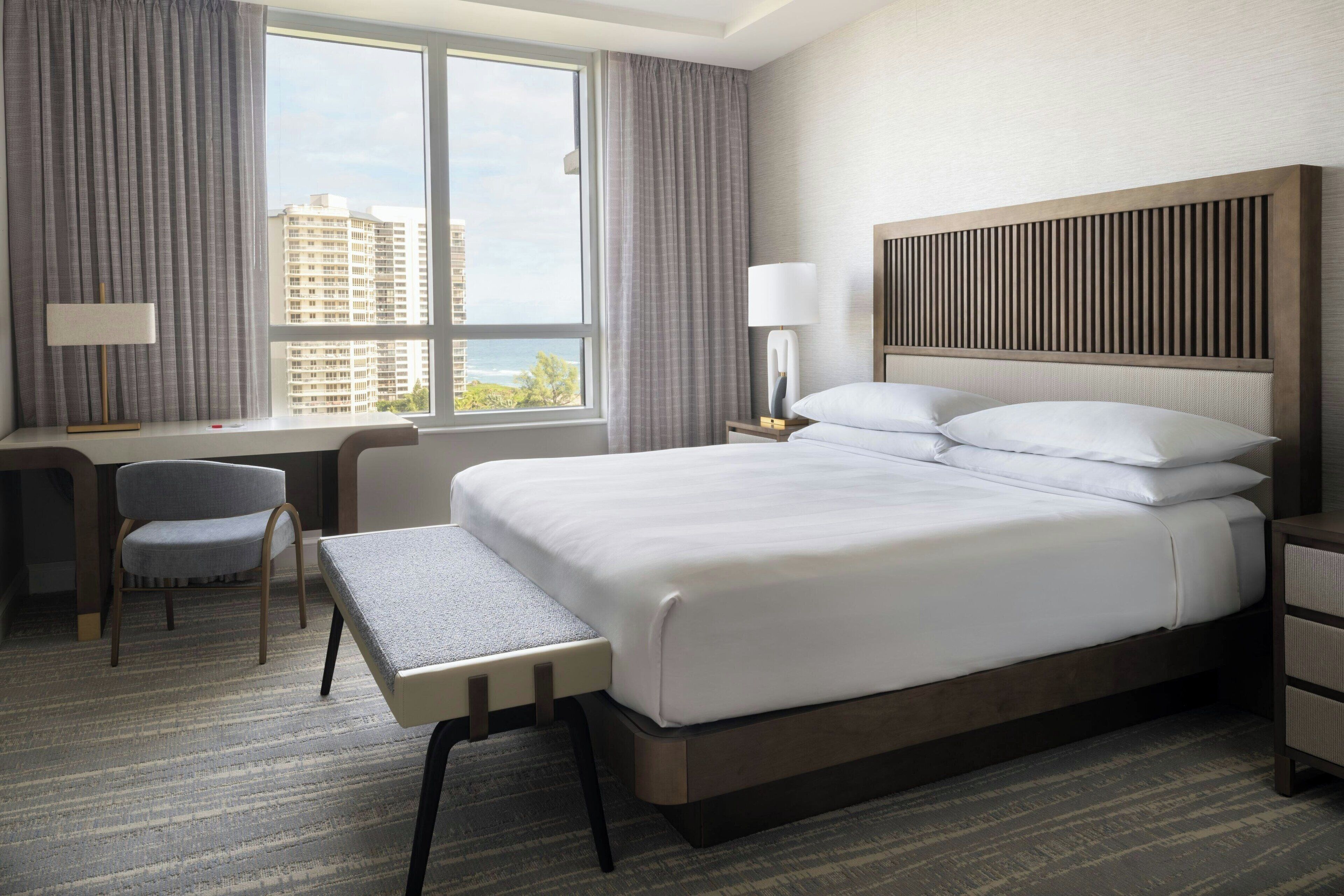 Habitación de hotel con una cama grande, un escritorio con silla, una mesita de noche con lámpara y vistas a edificios a través de la ventana.