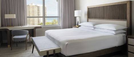 Habitación de hotel con una cama grande, un escritorio con silla, una mesita de noche con lámpara y vistas a edificios a través de la ventana.