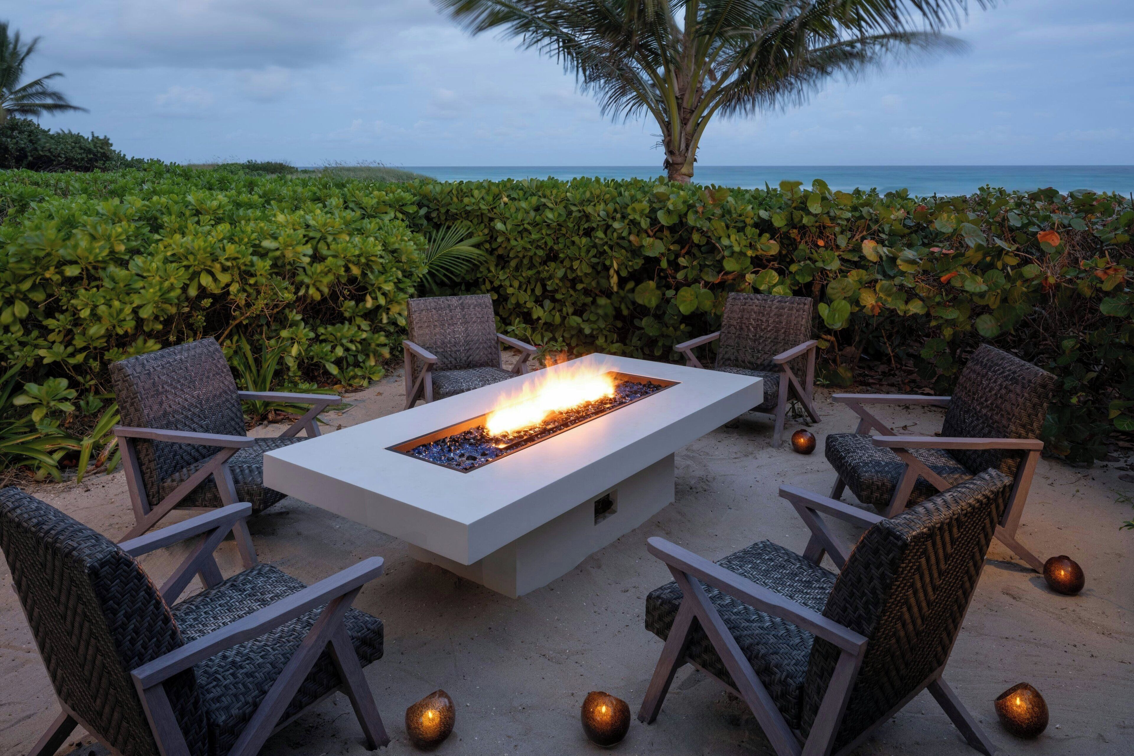 A fire pit table surrounded by wicker chairs, set on a sandy beach with a view of the ocean and a palm tree in the background.