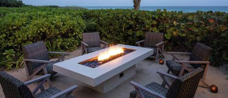 A fire pit table surrounded by wicker chairs, set on a sandy beach with a view of the ocean and a palm tree in the background.