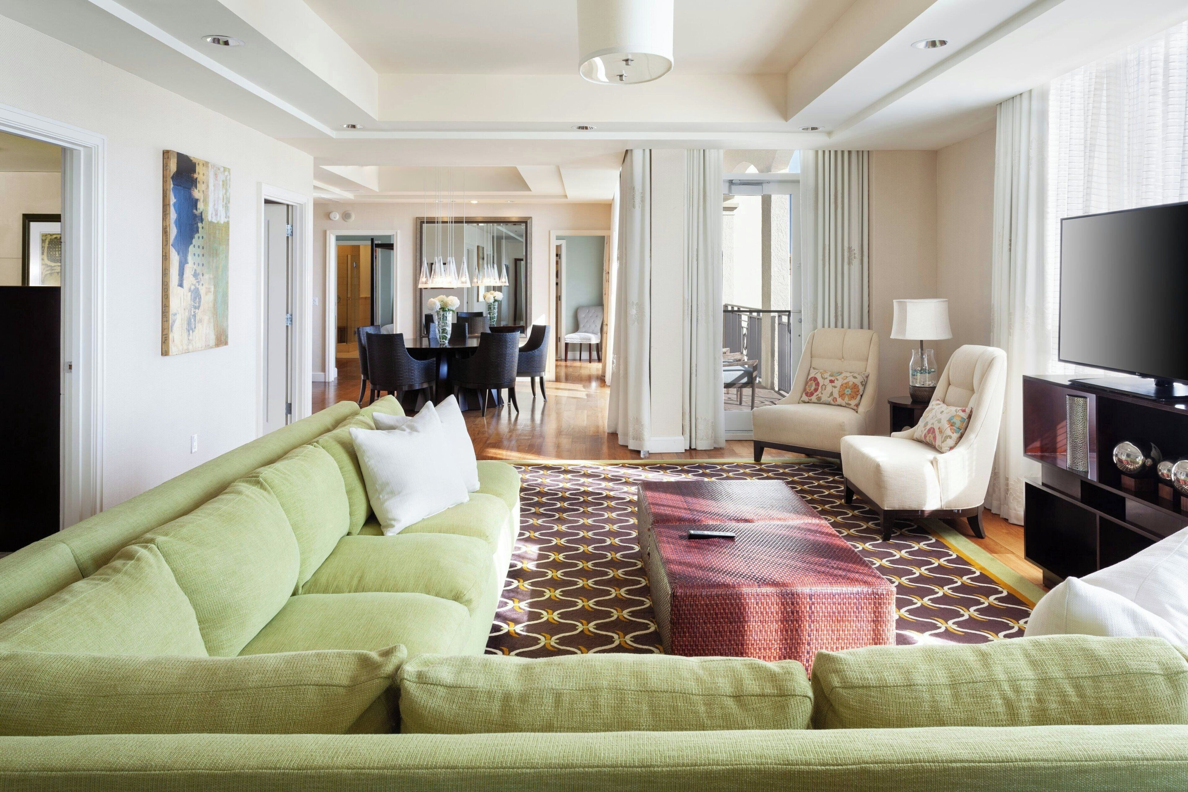 A modern living room with a green sofa, a patterned rug, a television, and a dining area in the background.