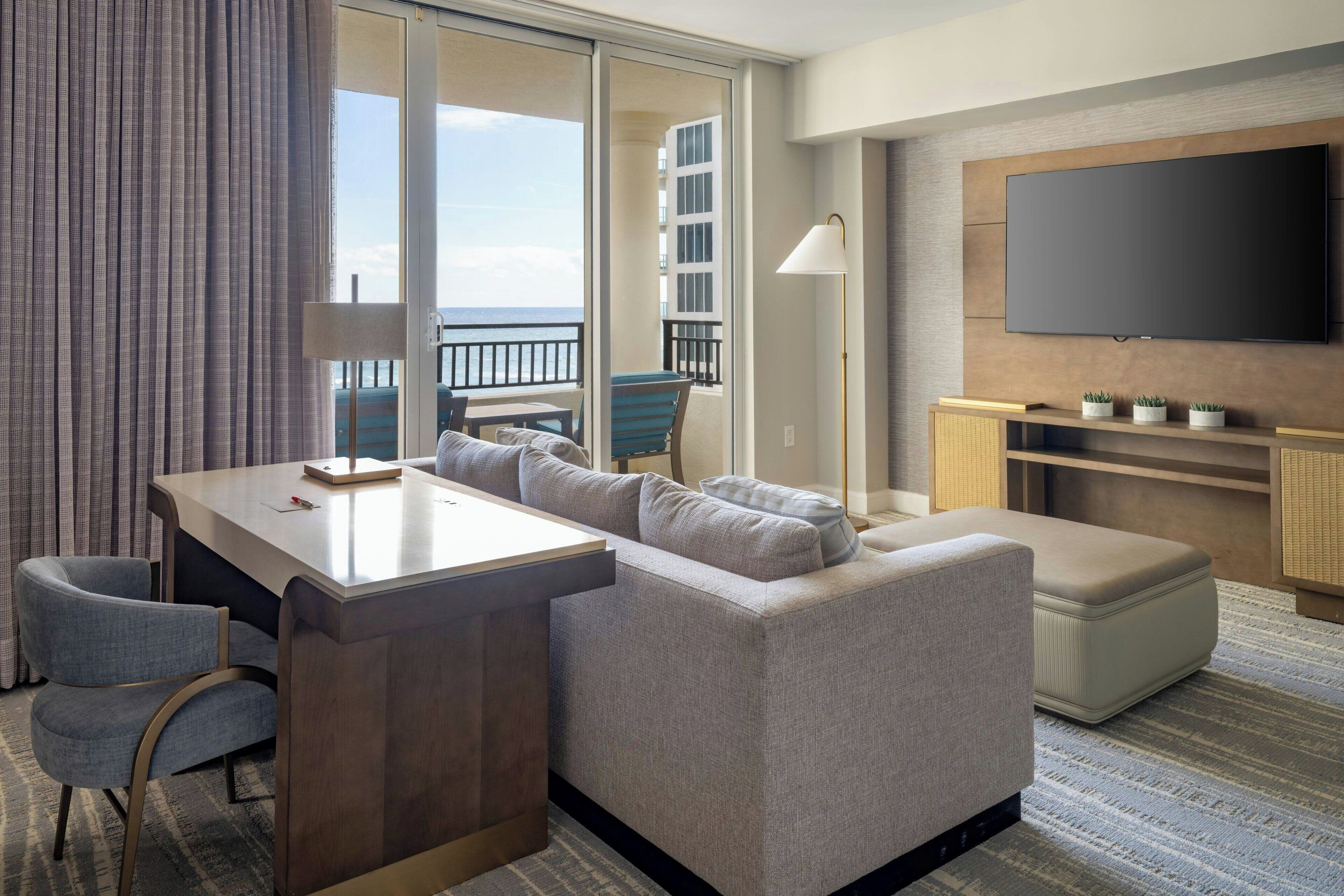 A hotel room with a large window, a sofa, a desk, and a flat-screen TV.