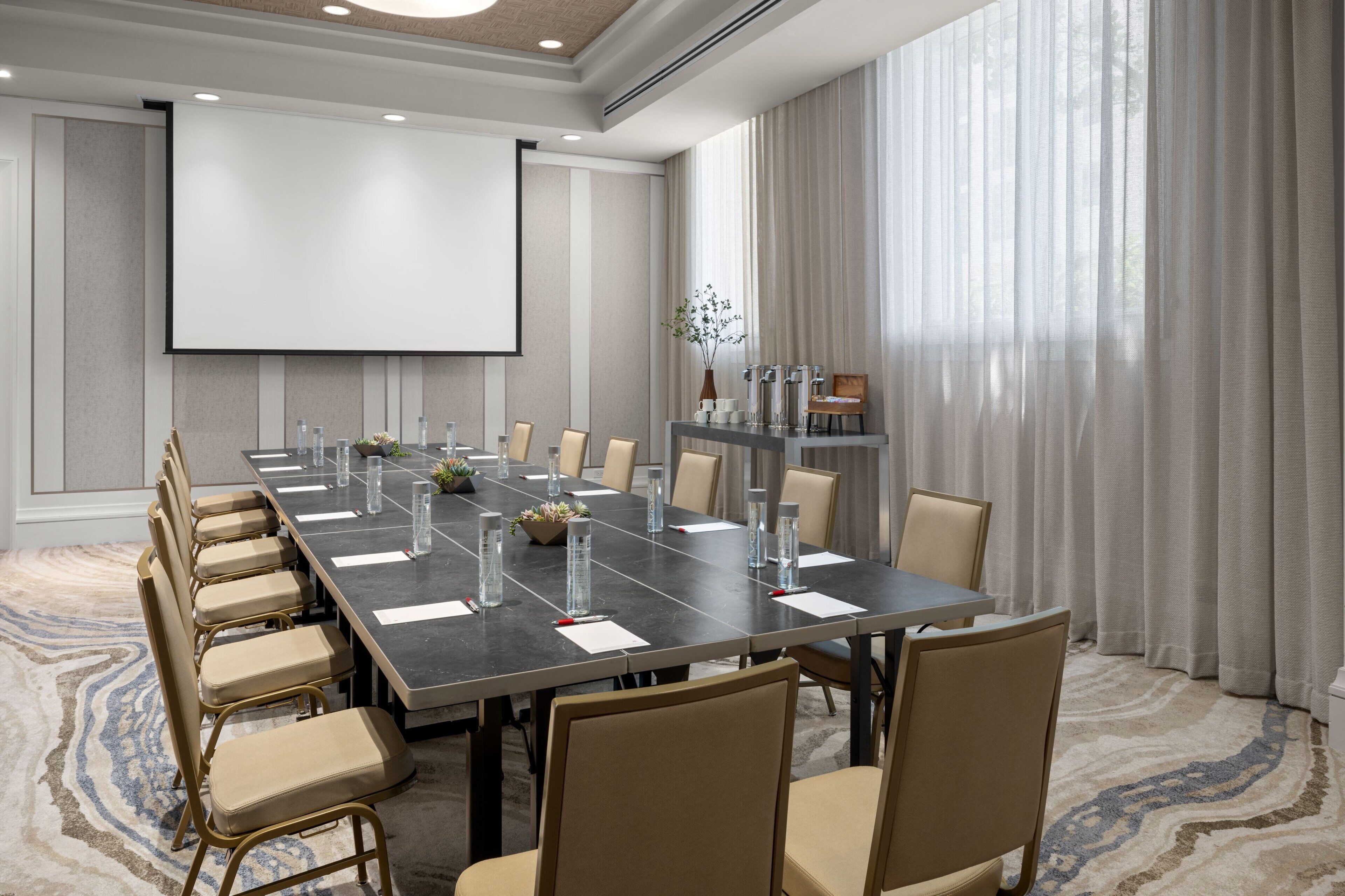 A conference room with a long table, chairs, a projector screen, and a large window with curtains.