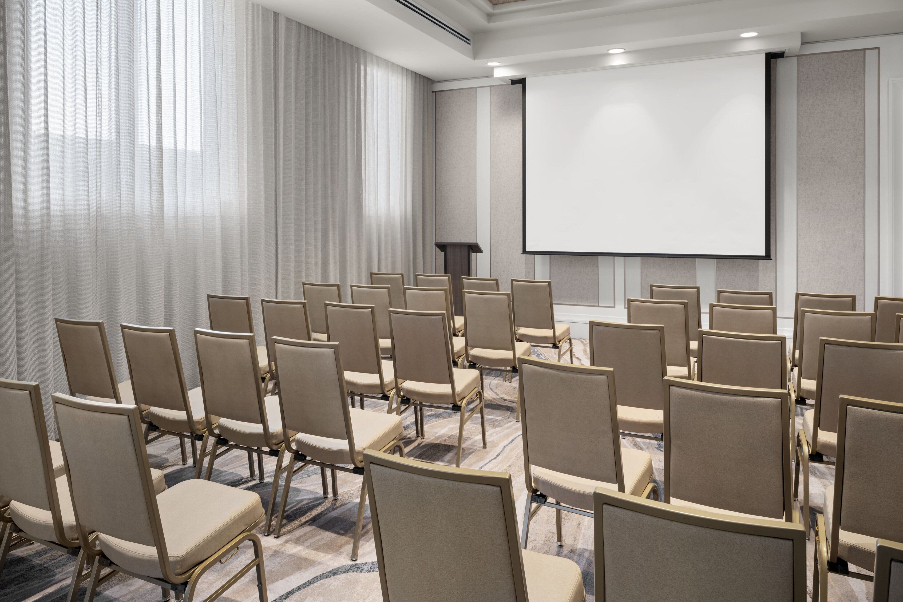 A conference room with rows of beige chairs, a projector screen, and a large window with sheer curtains.