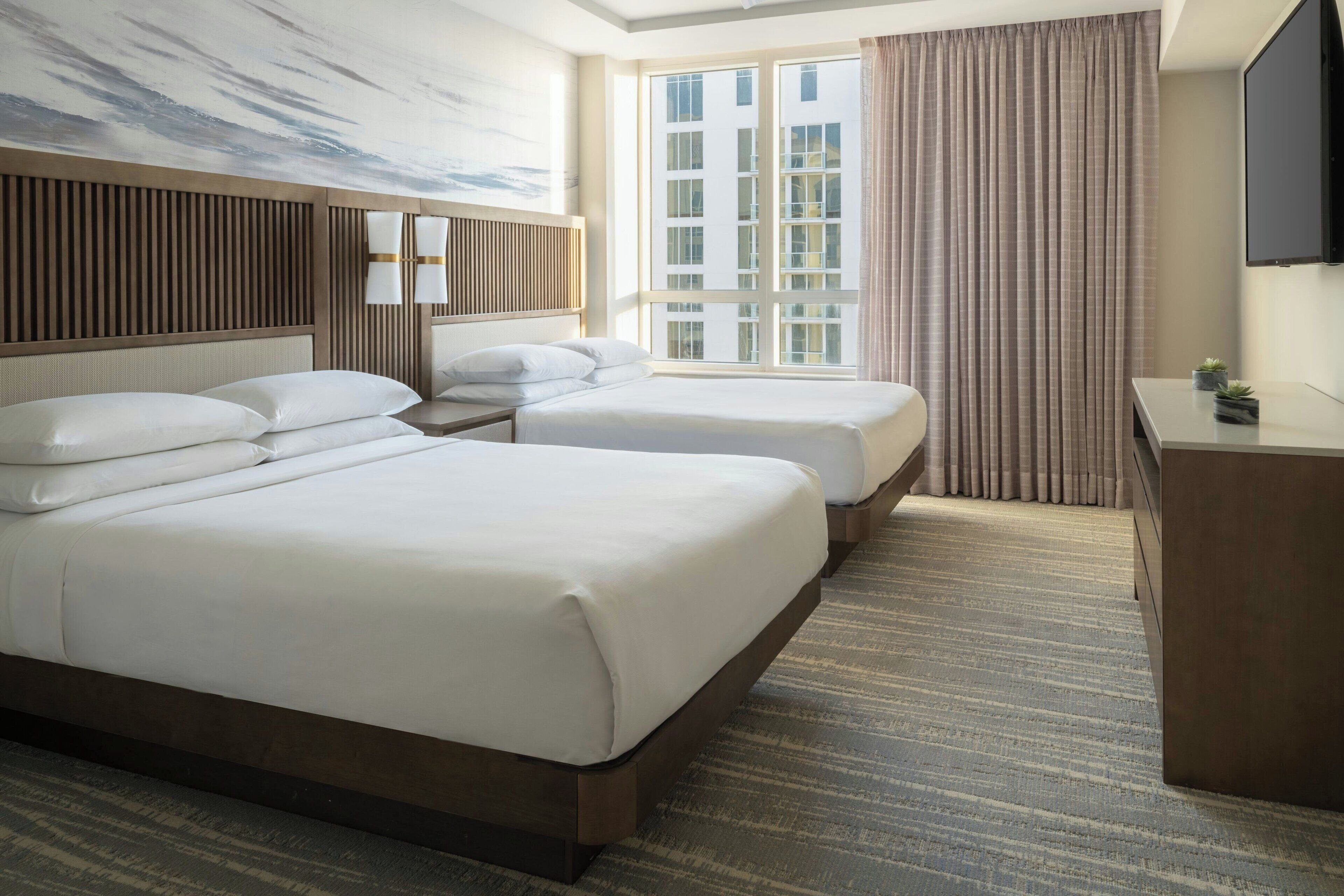 A hotel room with two beds, a desk, a TV, and a large window with curtains.