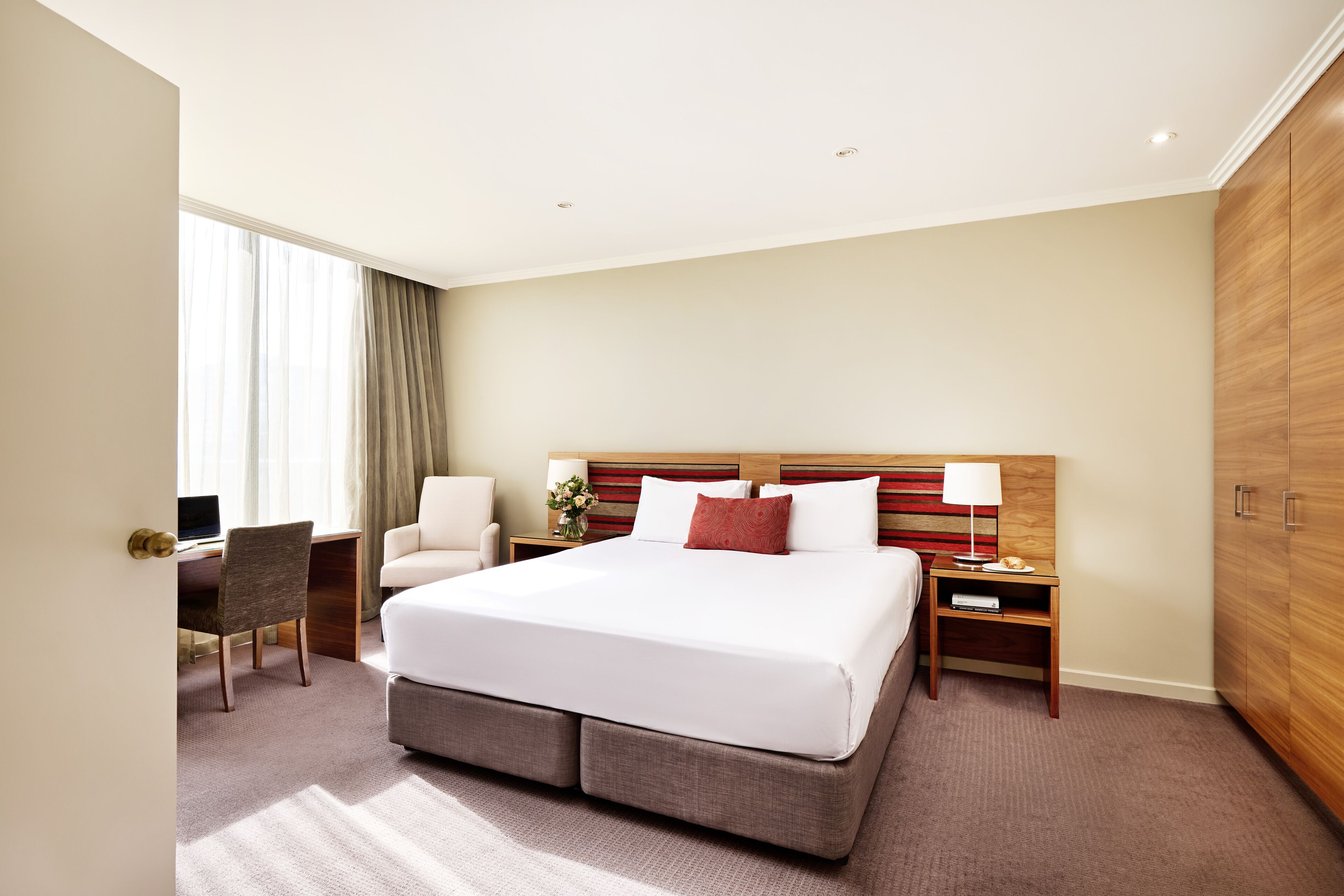 Quarto de hotel com uma cama grande, uma escrivaninha, uma cadeira, uma mesa de cabeceira e um guarda-roupa.