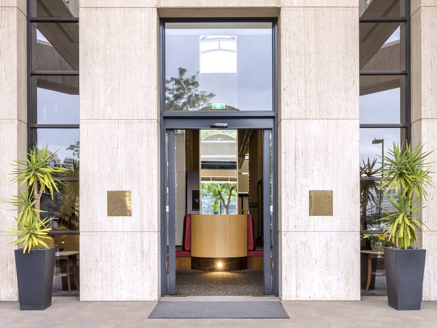 Una entrada moderna de edificio con puerta de vidrio, plantas grandes en macetas y una estructura de techo curva.