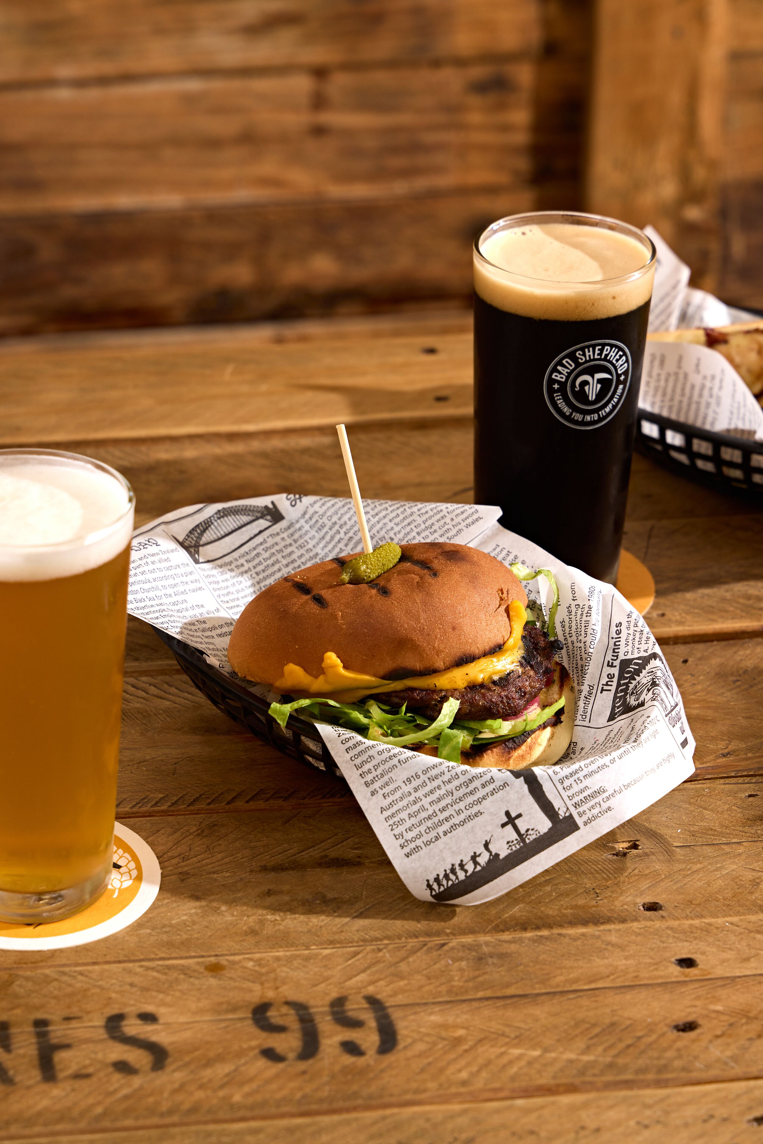 Una hamburguesa con papas fritas sobre un periódico, una jarra de cerveza y una taza negra con un logotipo, todo sobre una mesa de madera.