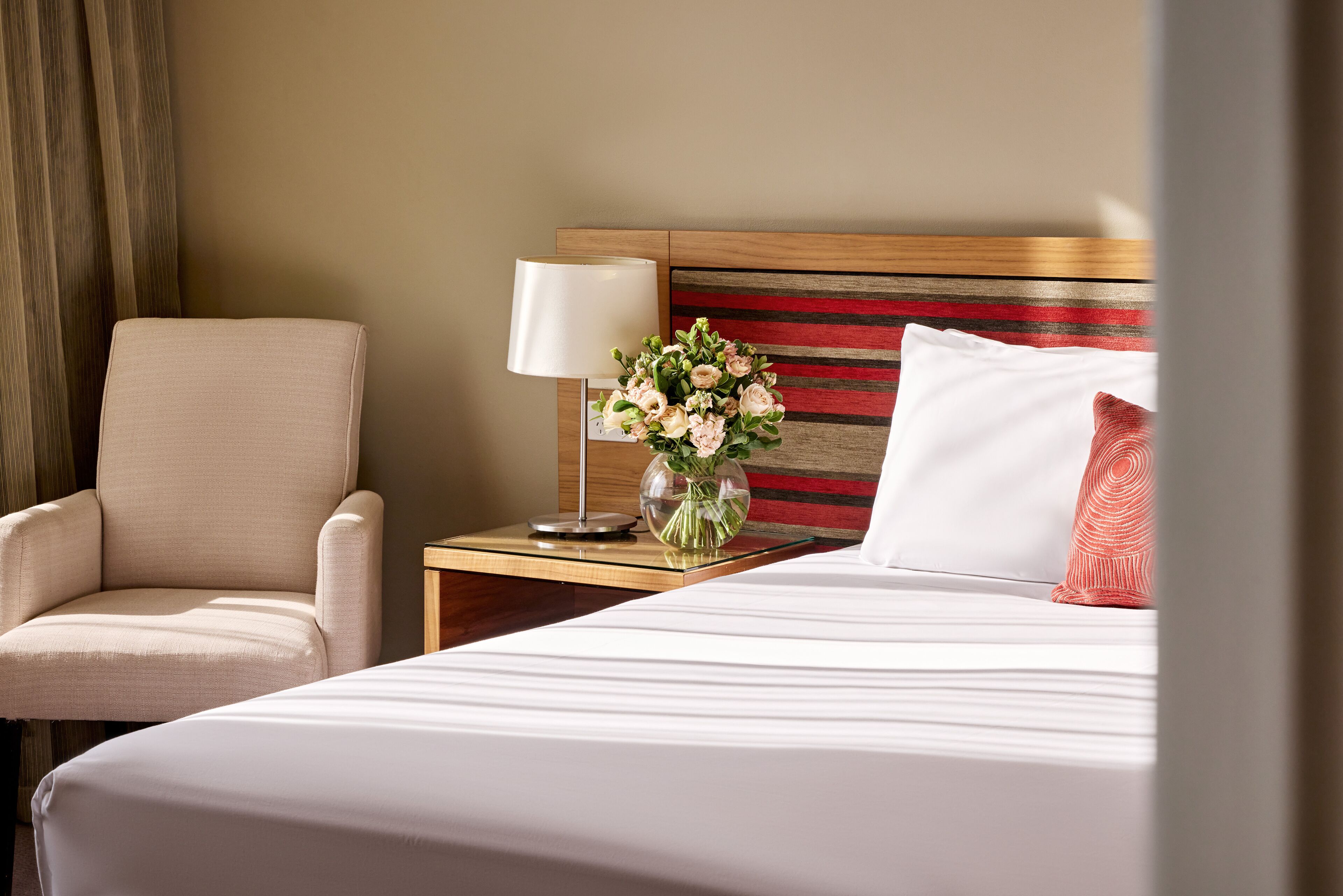 Una habitación de hotel con cama, una silla, una mesita de noche con lámpara y un jarrón con flores.