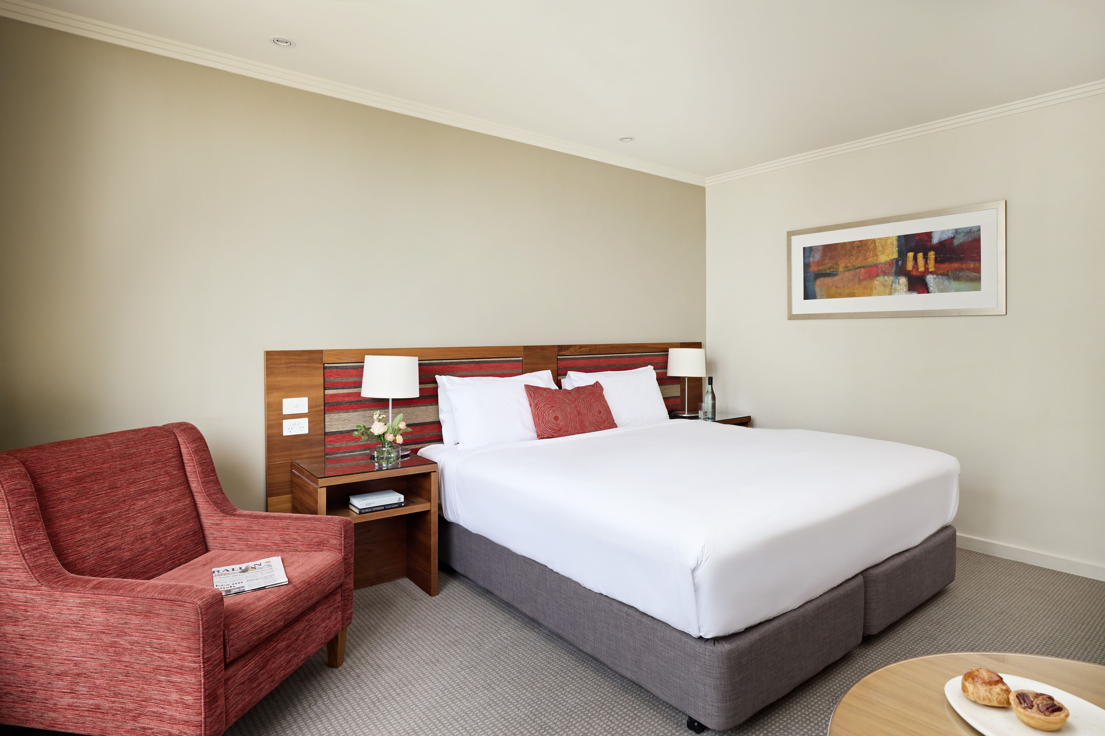A hotel room with a bed, a red armchair, a nightstand, a lamp, and a painting on the wall.