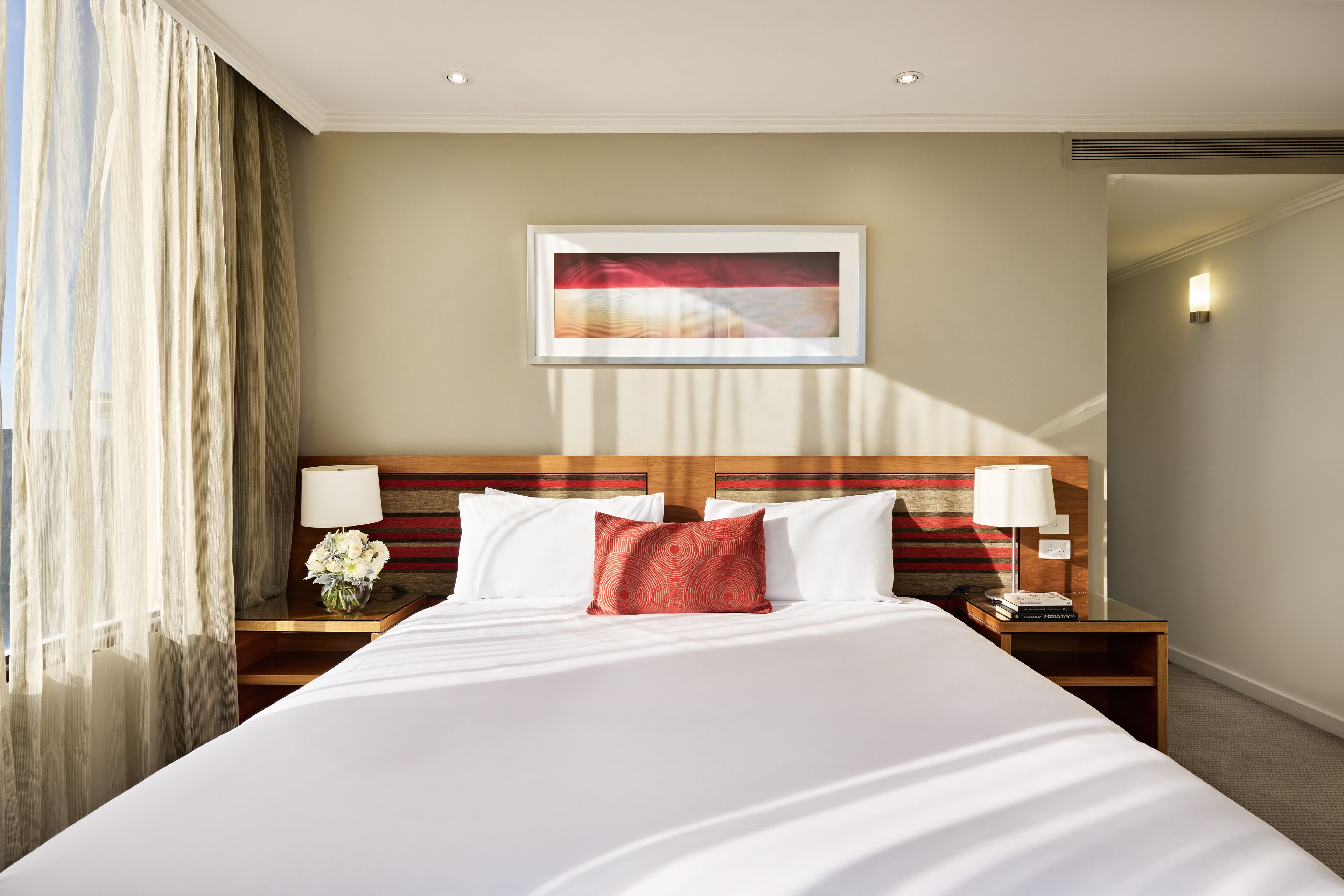 A hotel room with a large bed, two bedside tables with lamps, a framed artwork above the bed, and a view of the hallway.