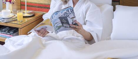 Une personne vêtue d’un peignoir blanc est assise sur un lit, un plateau rempli de divers aliments pour le petit-déjeuner devant elle, et elle est en train de lire un magazine.