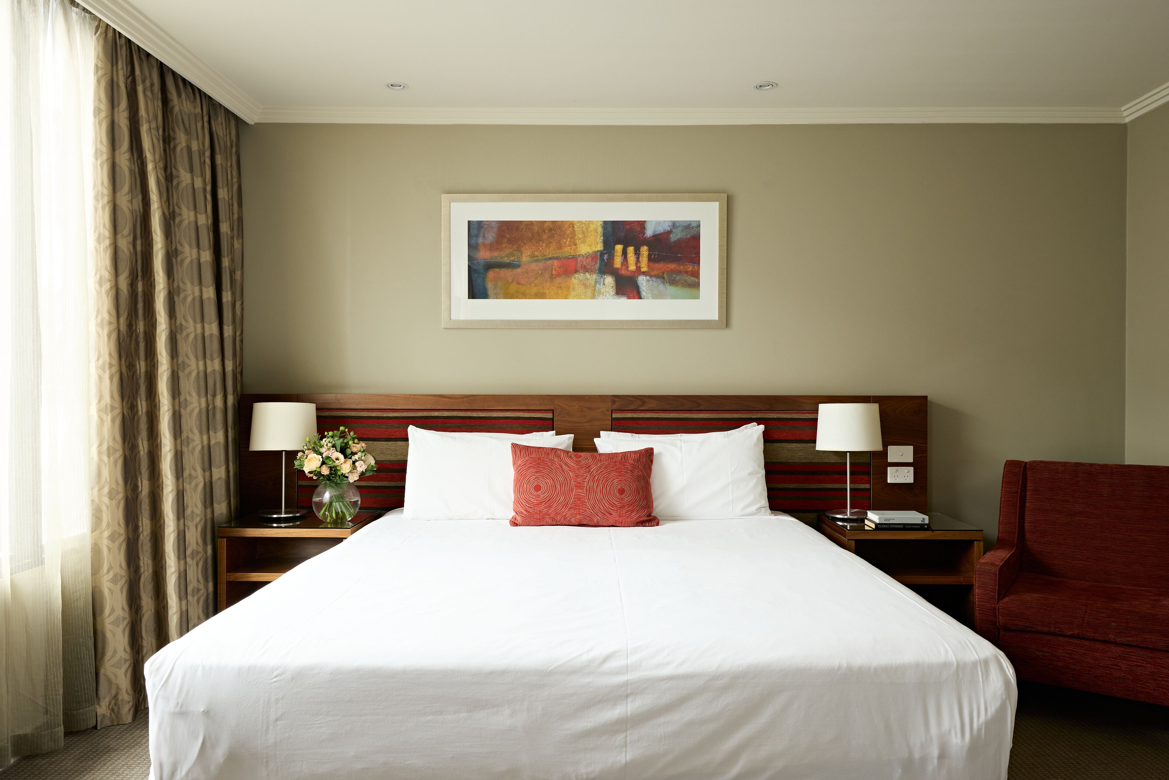 Habitación de hotel con una cama grande, una mesita de noche con lámpara, una silla roja y un cuadro en la pared.