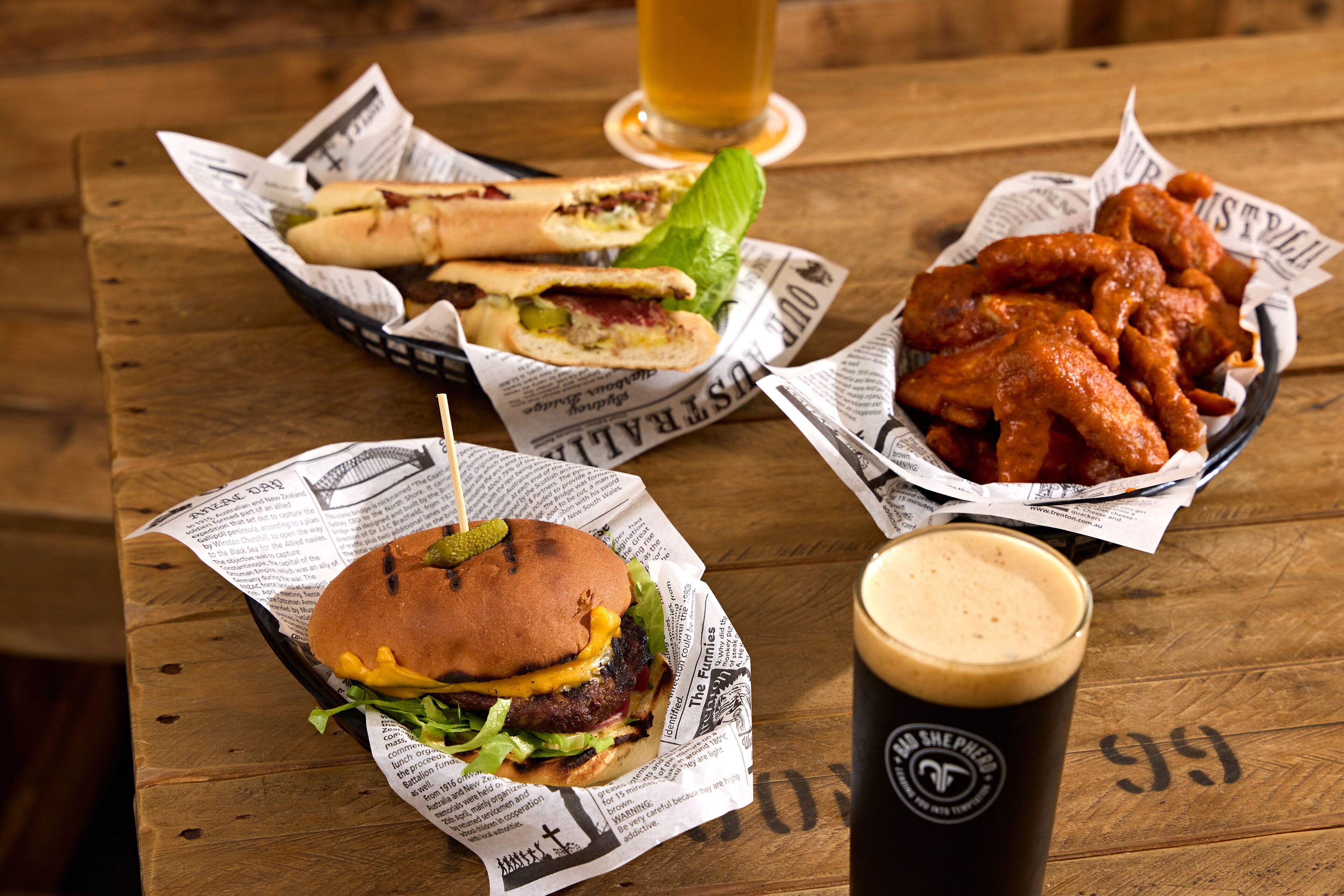 A wooden table with a burger, wings, and a drink on newspaper-lined trays.