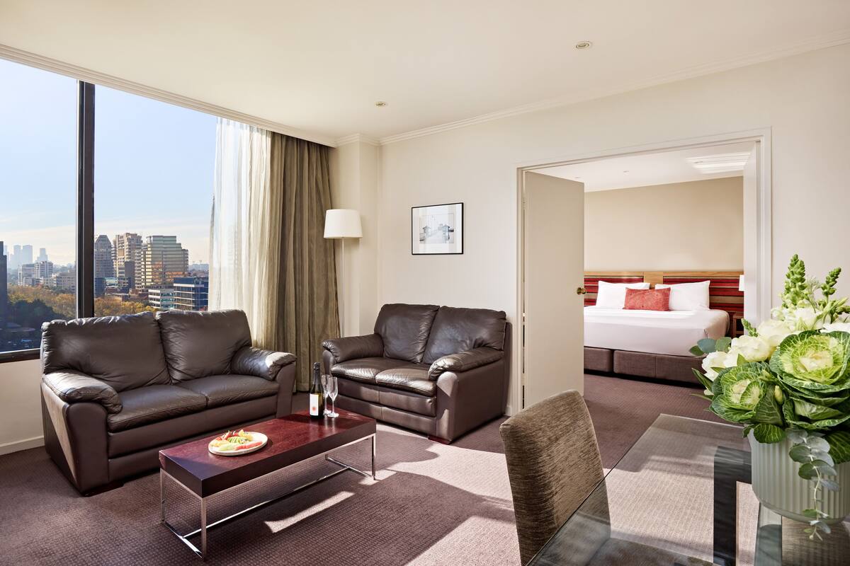 A hotel room with a city view, two leather sofas, a dining table, a bed, and a large window.