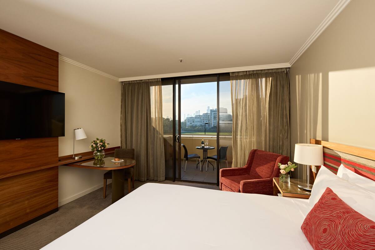 A hotel room with a large bed, a TV, a desk with a lamp, a chair, and a balcony with a view of the city.