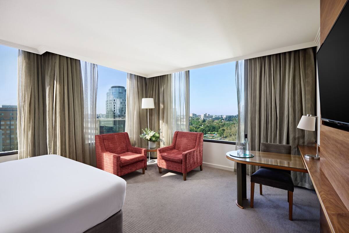 A hotel room with a large bed, two red armchairs, a small table with a lamp, a desk with a chair, and a TV.
